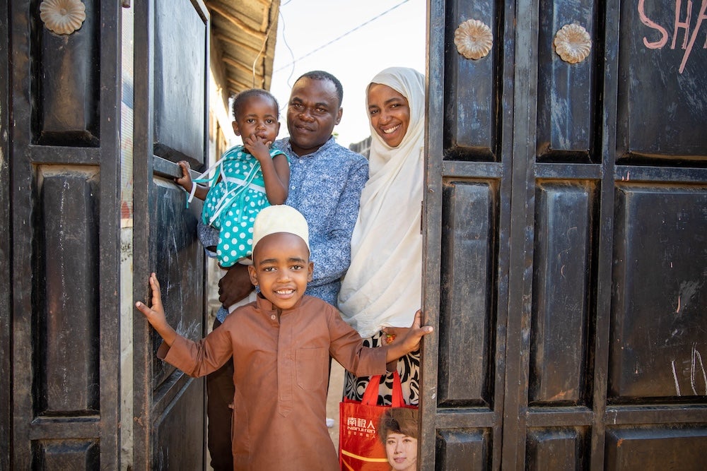 Une famille de quatre personnes dans l’encadrement de la porte de leur maison. Le père tient sa petite fille dans ses bras. La mère, qui porte un hijab blanc, sourit largement. Au premier plan, leur fils tient la porte ouverte.