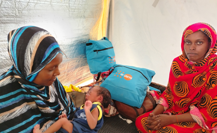  Una mujer vestida de rojo sonríe ligeramente a la cámara sentada en una tienda de campaña, con bolsas del UNFPA visibles detrás de ella. Otra mujer en rayas azules y negras sostiene a un bebé que llora.]