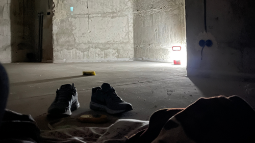 Une paire de chaussures d’homme près d’un tas de couvertures, dans un bunker souterrain. Une lampe éclaire l’angle de murs en béton